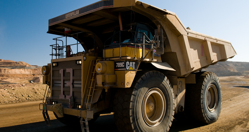Large yellow mining dump truck driving on a dirt road at a quarry with a clear blue sky in background.